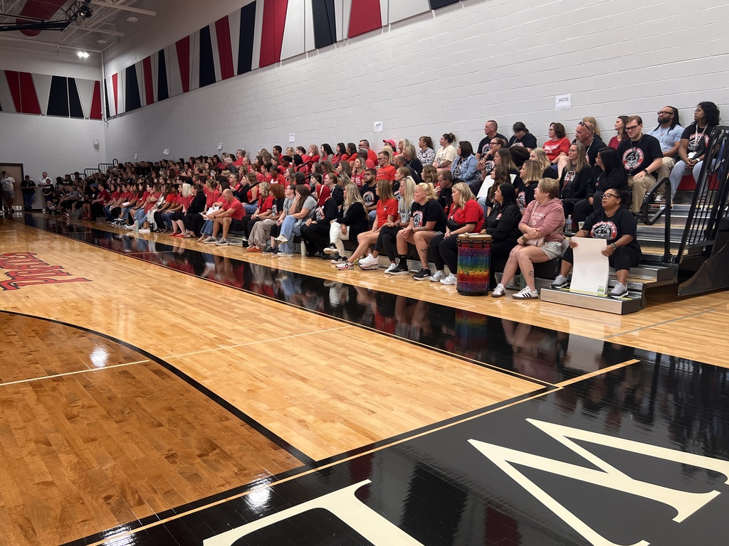 A crowd gathers on bleachers in a gym, with a basketball court below and a white wall with banners.
