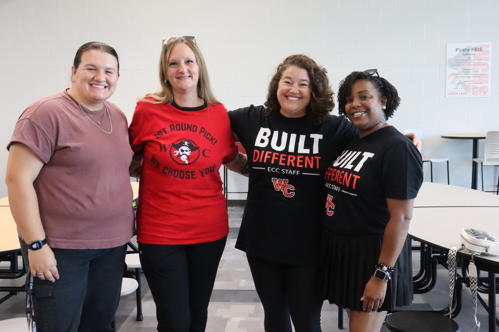 Four women stand in a classroom, smiling. They wear various colored t-shirts with "Built Different" text. Tables and chairs are in the background.