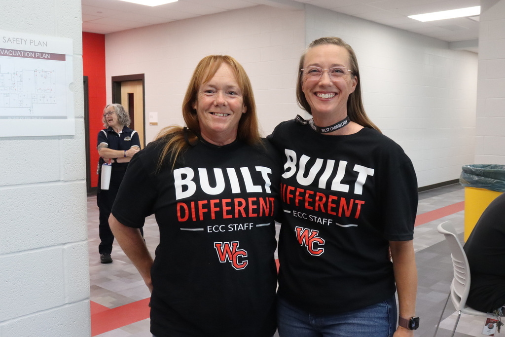 Two women stand next to each other in a hallway, wearing matching black t-shirts that say "Built Different."