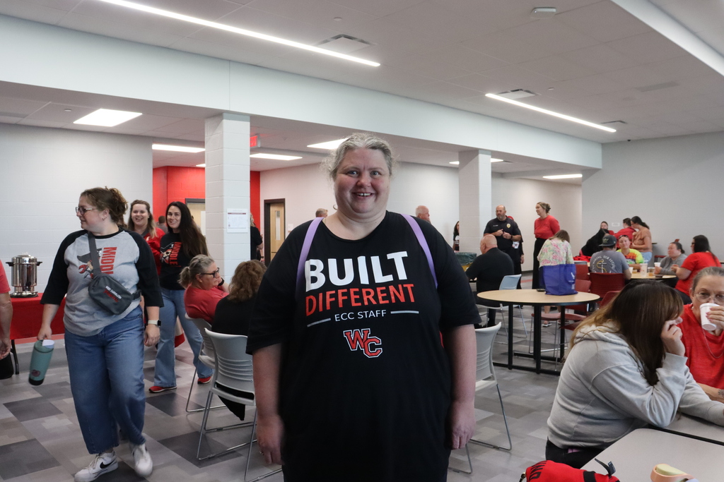 A smiling woman stands in a room with other people, tables, chairs, and a shirt that reads "BUILT DIFFERENT."