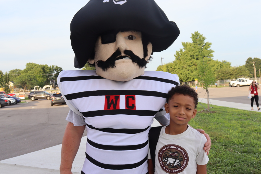 A man in a pirate mascot costume stands with a young boy in a schoolyard.
