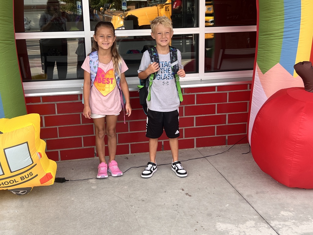 Two kids stand in front of a colorful wall. A girl wears a pink shirt and pink shoes. A boy wears a gray shirt, black shorts, and sneakers.
