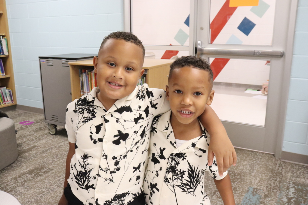 Two boys, both wearing white and black patterned shirts, are smiling at the camera. Behind them is a bookshelf, a door, and a colorful wall.