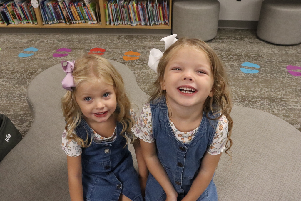 Two girls in blue overalls, one with a pink bow, sit on a curved surface, smiling. Behind them, shelves hold books.