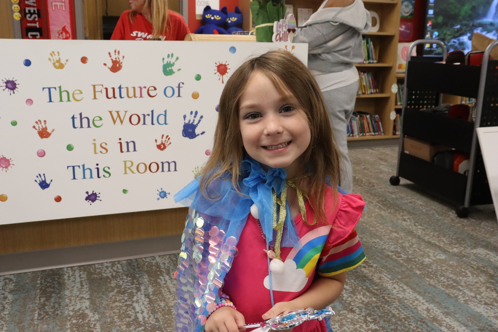 Girl wearing colorful clothing and a cape with sequins stands in front of a sign with handprints.