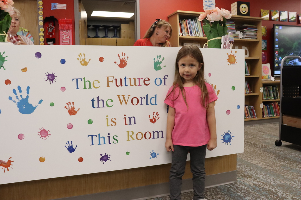 A young girl in a pink shirt stands by a banner that reads "The Future of the World is in This Room."
