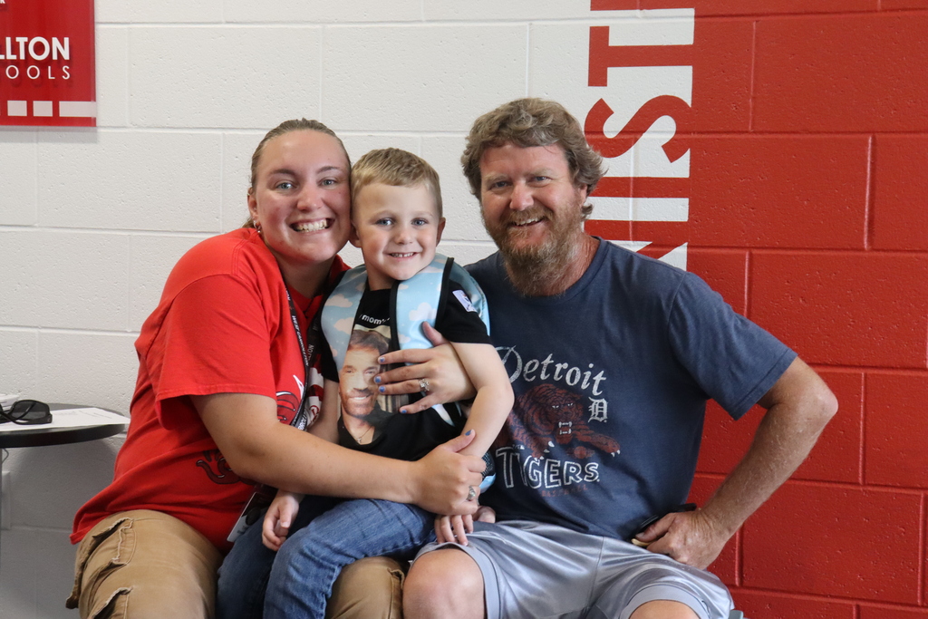 Three people smiling: a woman in red, a man in blue, and a child in black, all seated in front of a red wall.