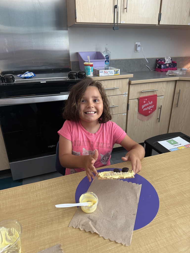 A young girl sits at a table with a graham cracker, frosting, and a paper towel