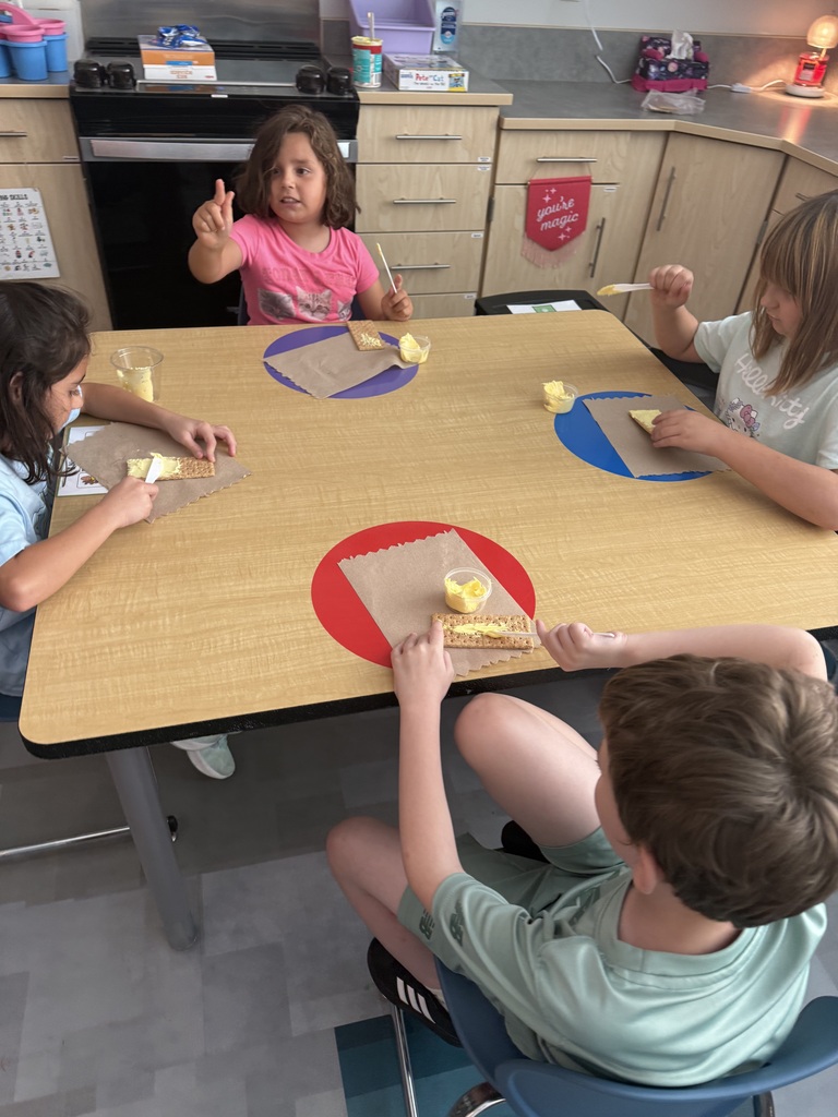 Four children sit at a kitchen table, each with a graham cracker, frosting, and paper towel in front of them.