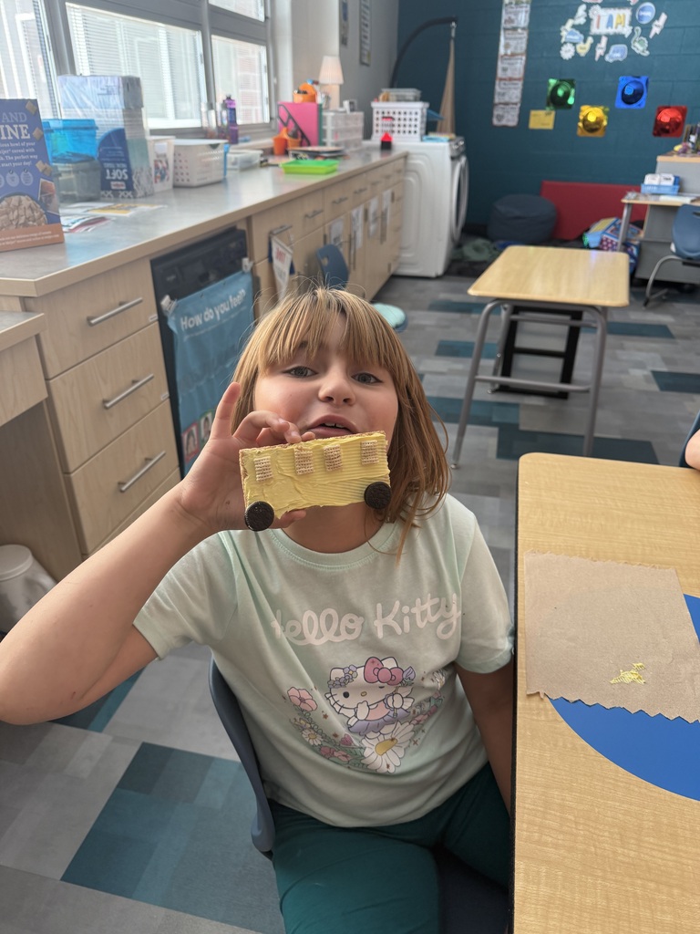 A child holds a graham cracker made to look like a school bus in a classroom, with tables and chairs in the background.