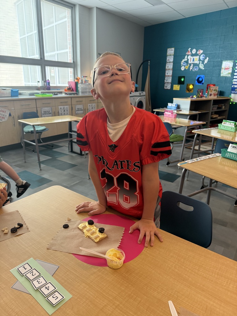 A child in a red jersey stands at a table, gazing upward, with a graham cracker and frosting on a paper towel..