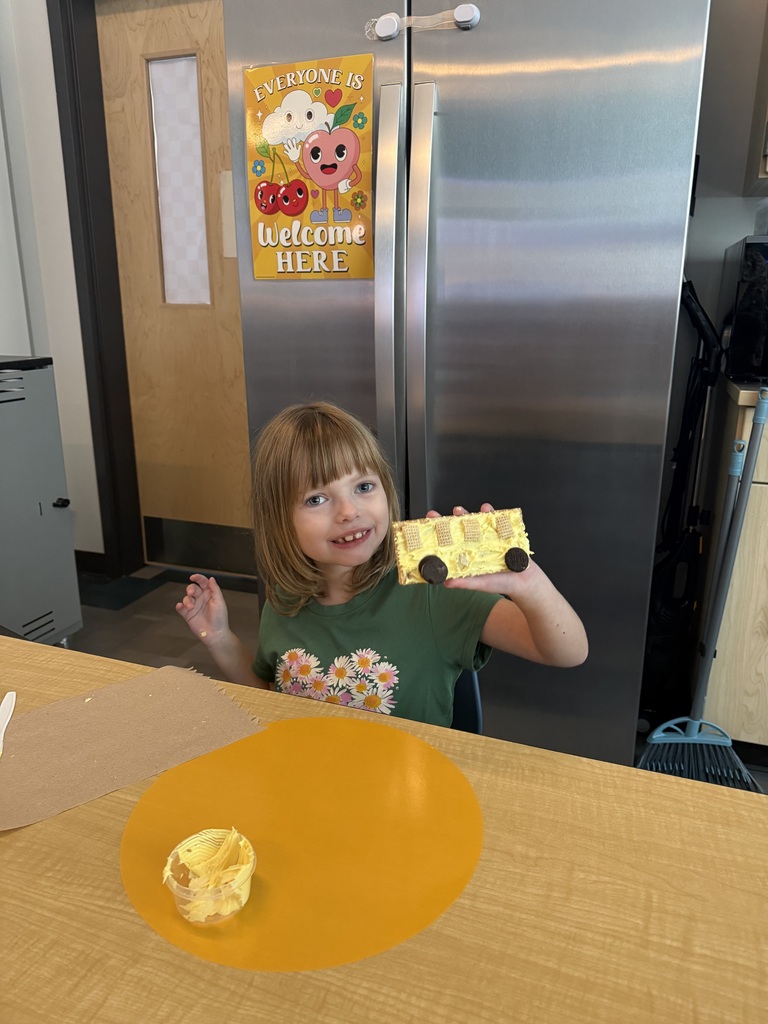 Young girl sitting at a table with a graham cracker made to look like a bus, against a backdrop of a refrigerator.