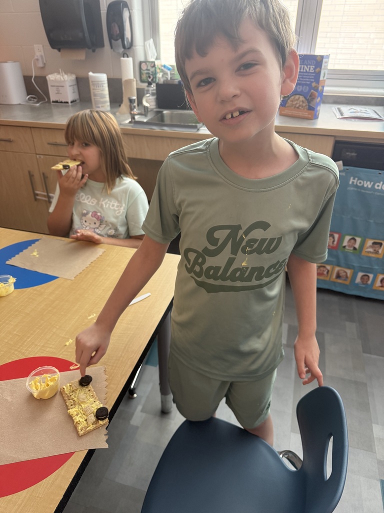 A boy in a green "New Balance" shirt stands near a table with food. Behind him, a girl eats from a plate.