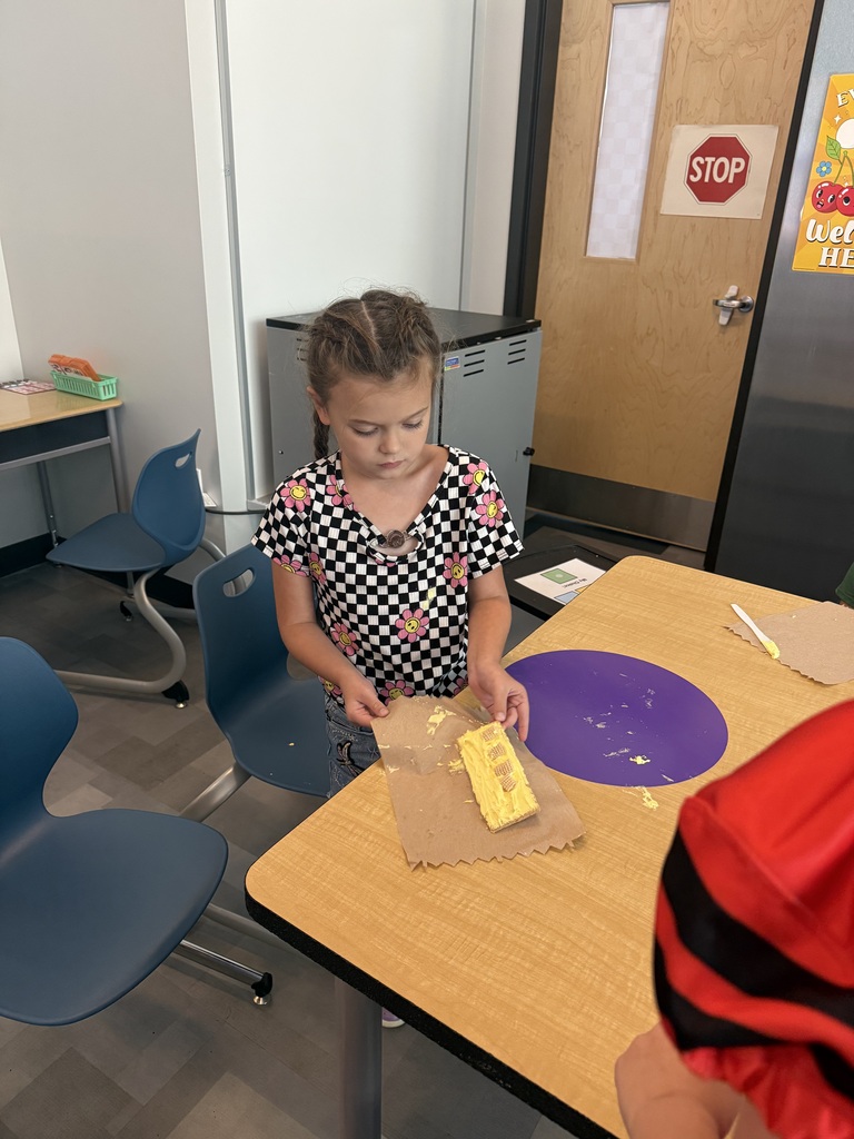 A young girl stands at a table, holding a graham cracker and paper towel, with another person partially visible.