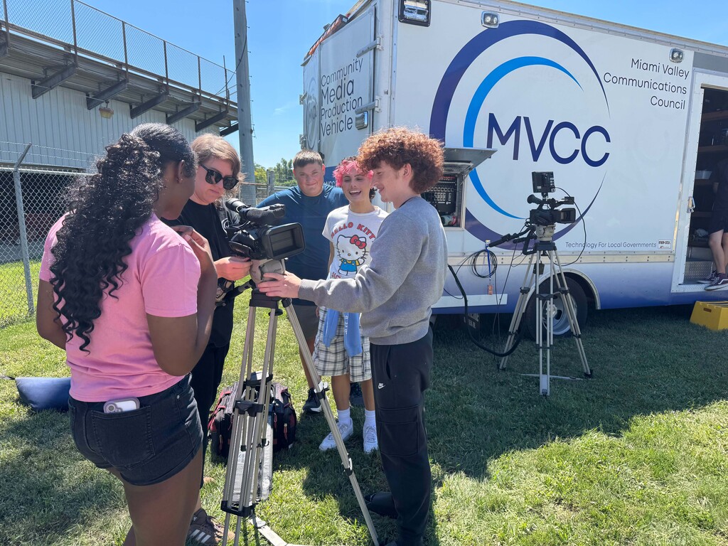 Several individuals are gathered on grass, operating camera equipment. A van with the text MVCC is visible in the background.