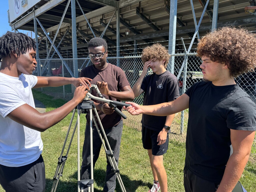 Four young men gather around a tripod, on a grassy field with a stadium in the background.