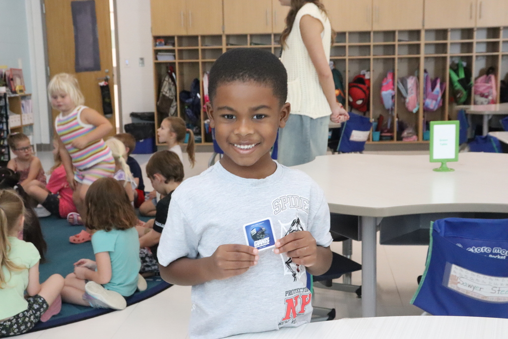 A smiling young boy holds a card in a classroom. Behind him, children sit on the floor, while two people stand.