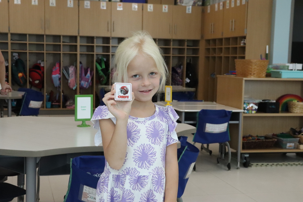 Blonde-haired child in a purple floral dress holds up a card.