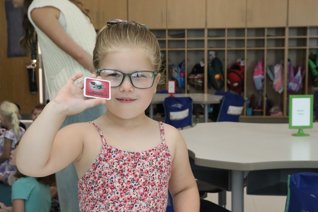 A young girl wearing glasses holds up a red card, smiling. Behind her, a classroom is visible with tables, chairs, and shelves.