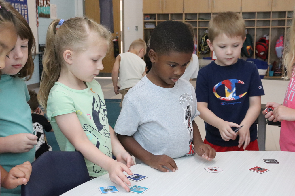 Several children, wearing casual clothing, are playing with cards on a table in a classroom.