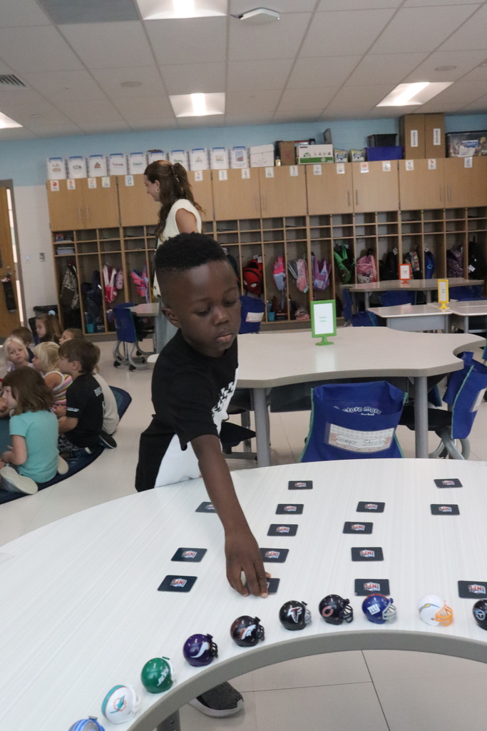 A child reaches for a card on a white table in a classroom with other students and a teacher.