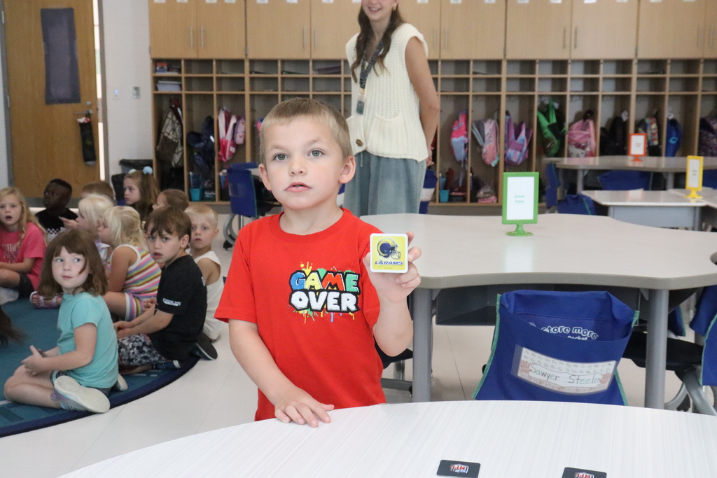 Child standing at a table, holding a small yellow card with text, while other children sit on the floor.