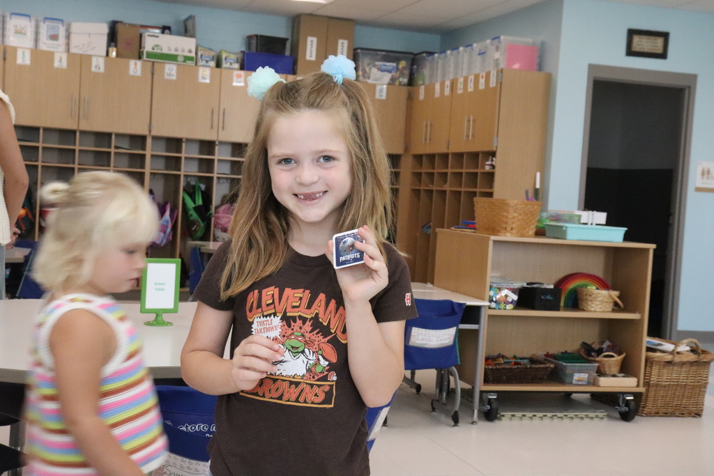 Two young girls, one holding a card, stand in a classroom with shelves, a basket, and a white desk.