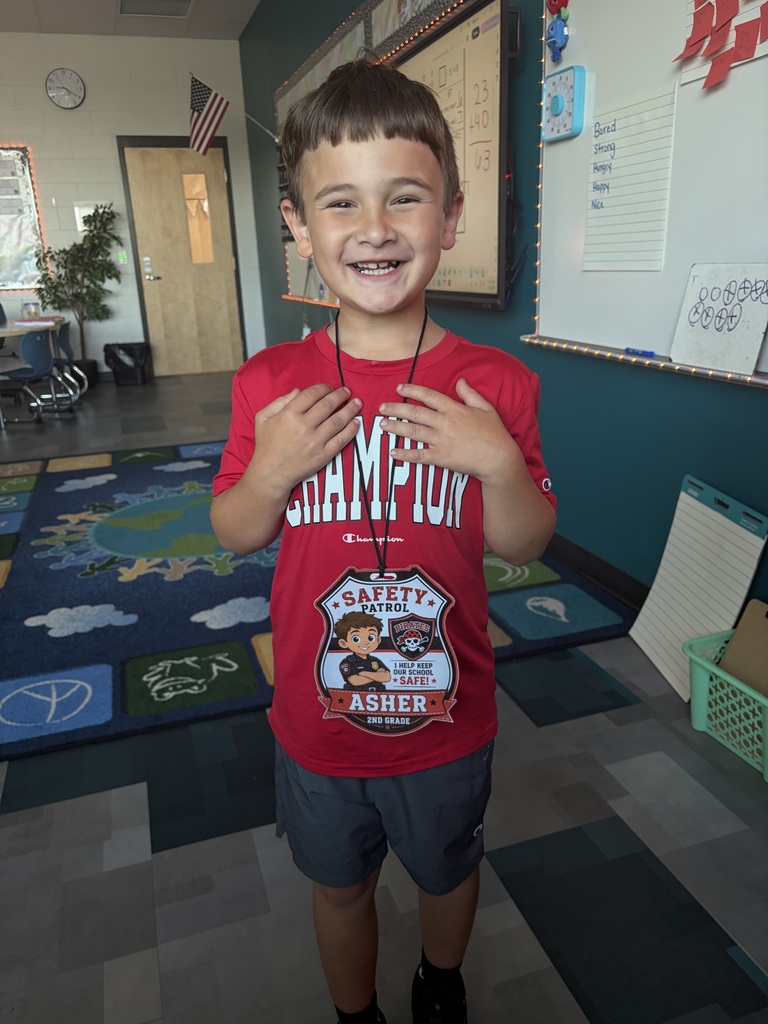 A boy stands in a classroom, smiling and holding a badge around his neck. Behind him are various classroom elements, including a bulletin board.