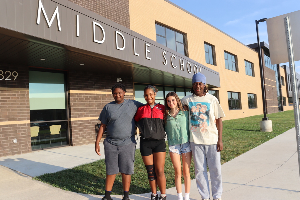 four students stand outside of a Middle School building with brick walls and windows in the background