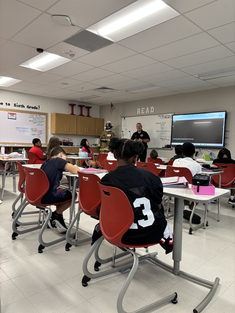 Classroom with students seated at desks. A person stands at a whiteboard. Monitor on the wall, tables, chairs, and ceiling lights.