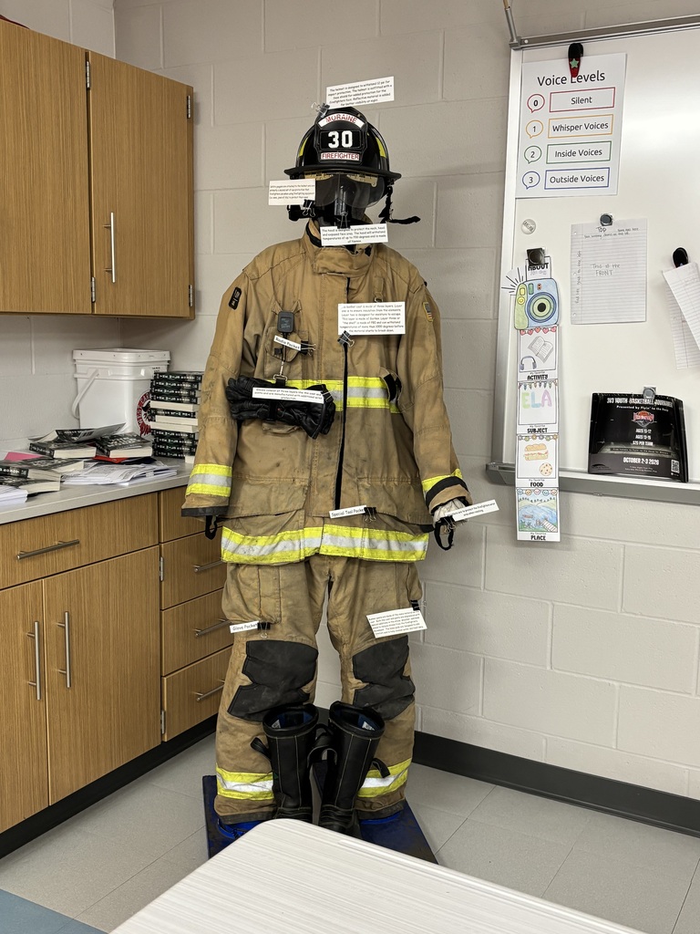 A mannequin dressed in a firefighter's uniform stands in a room with a whiteboard and wooden cabinets.