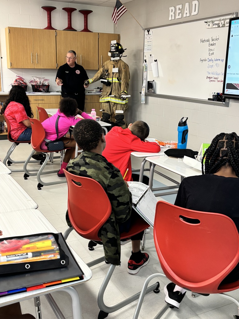 Classroom with a fireman and a fireman's outfit at the front. Students seated in red chairs.