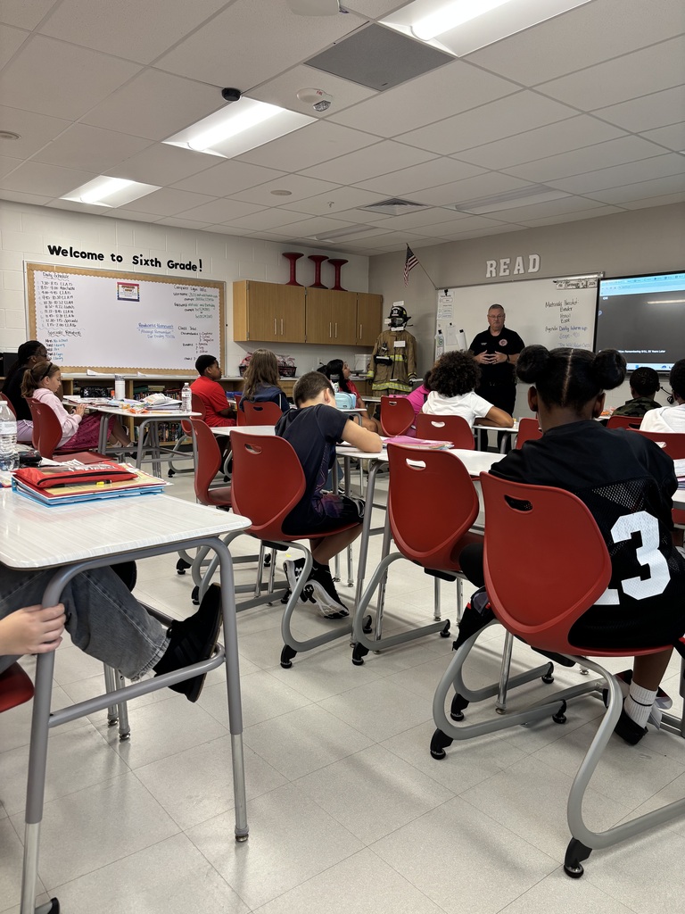 Classroom with desks and chairs. A person stands at a whiteboard. Students are seated in rows.