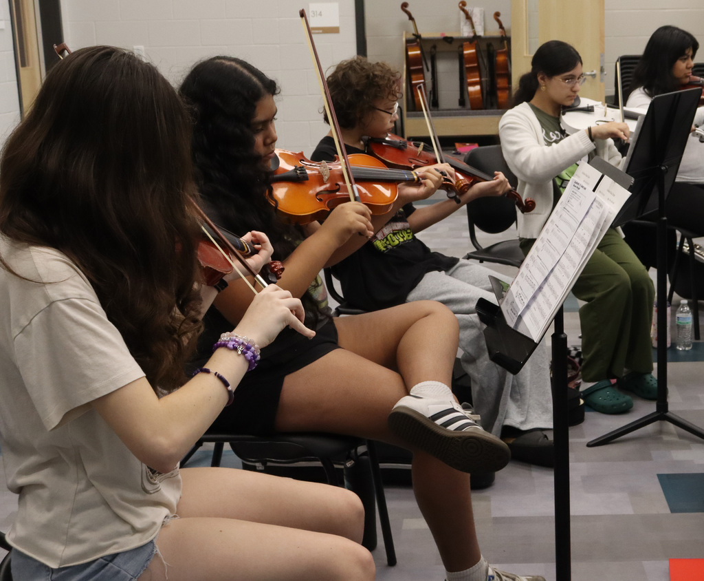 Group of young musicians playing violins in a classroom setting with music stands and sheet music.