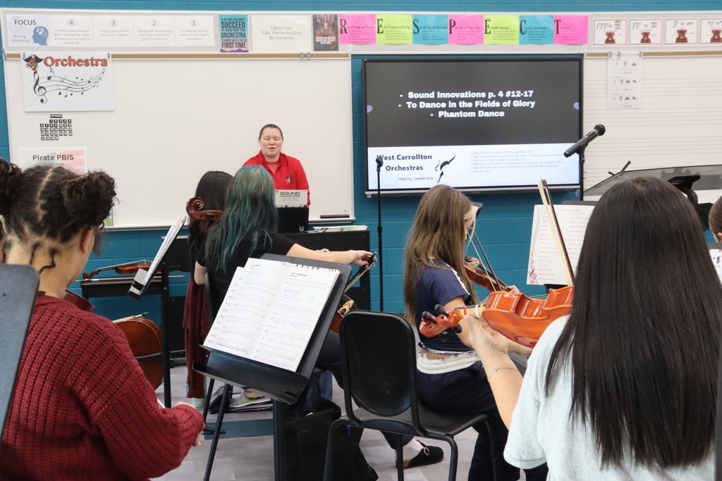 Classroom with students seated, holding violins and bows. A teacher stands by a screen showing text.