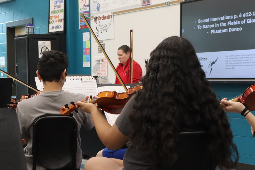 A classroom with a teacher, a projector, and students playing violins. Long-haired student in foreground.