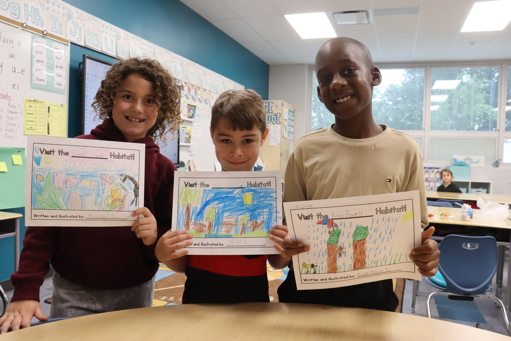 Three children hold up drawings of landscapes, smiling and standing in a classroom. One has a red shirt, another a maroon shirt, and the third wears tan.