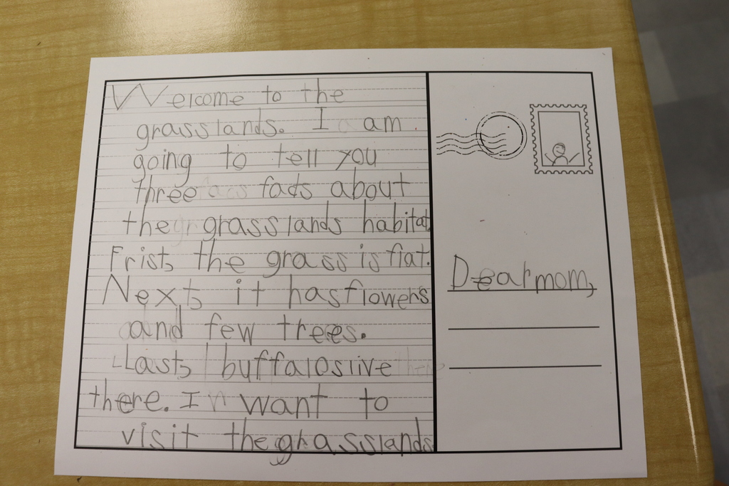 A handwritten page displays text about grasslands, mentioning grass, flowers, and trees. The page is on a wooden surface.
