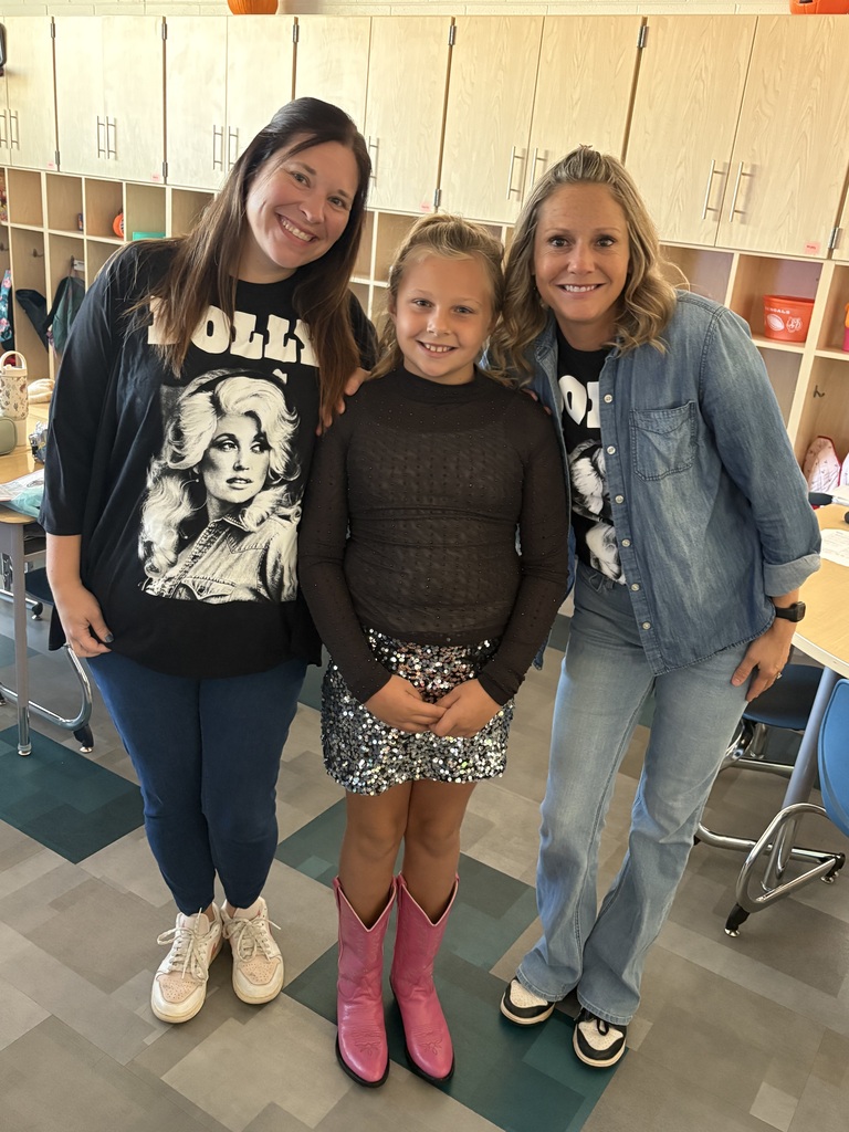 Two women and a child stand in a classroom; one wears pink boots, one wears a denim jacket. Behind them, wooden cabinets and a patterned floor.