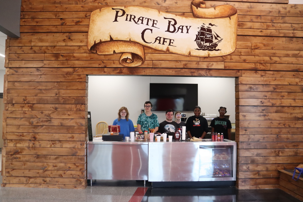 Five people stand behind a counter that has cups and bottles on it. There is a wood paneled wall with the sign that says "Pirate Bay Cafe" above them.