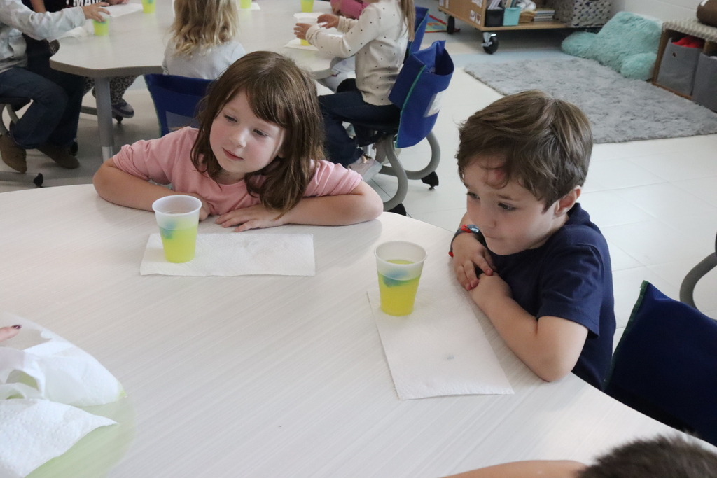Two children sitting at a table looking at a cup with yellow liquid and a blue ice cube.