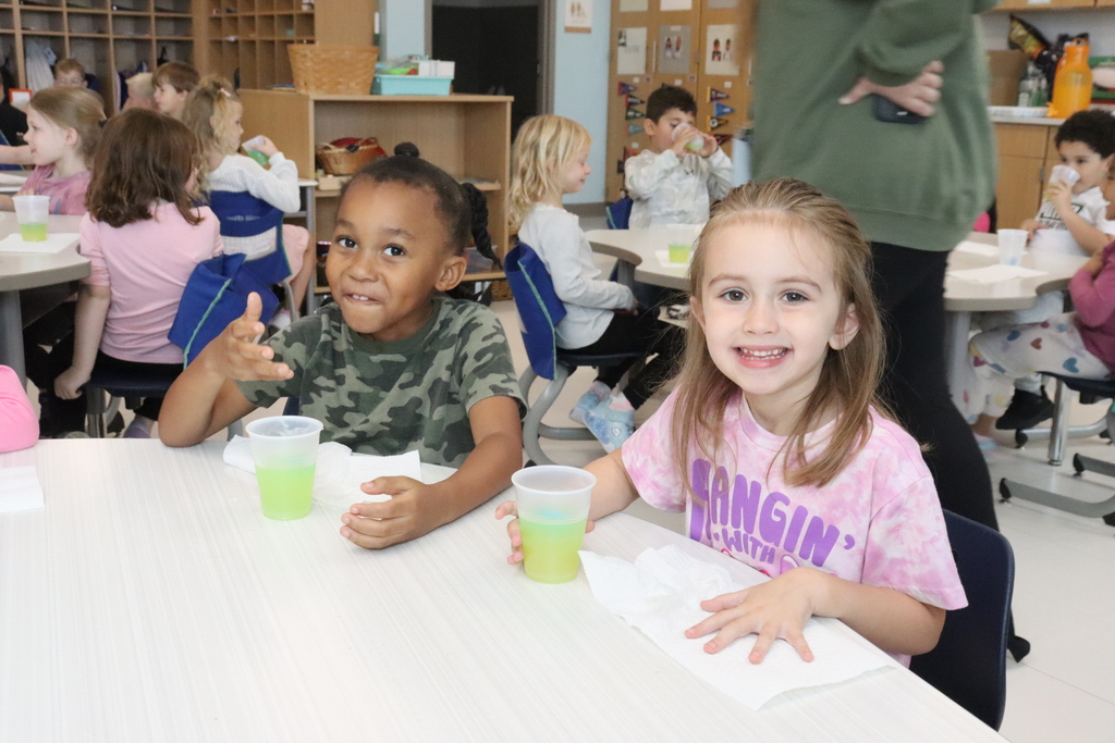 Children seated at tables with green drinks. Two smiling girls sit next to each other. One is wearing a pink shirt and the other is wearing a camo shirt.