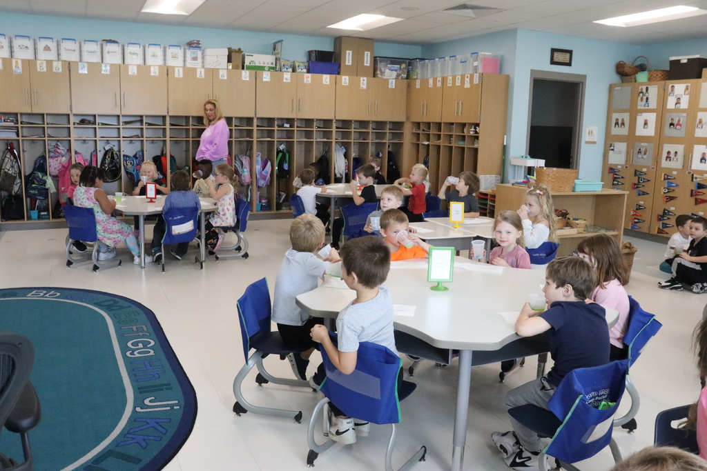 Classroom with young children seated at tables, a teacher in pink stands in the background with wooden cabinets, and a blue rug is on the floor.