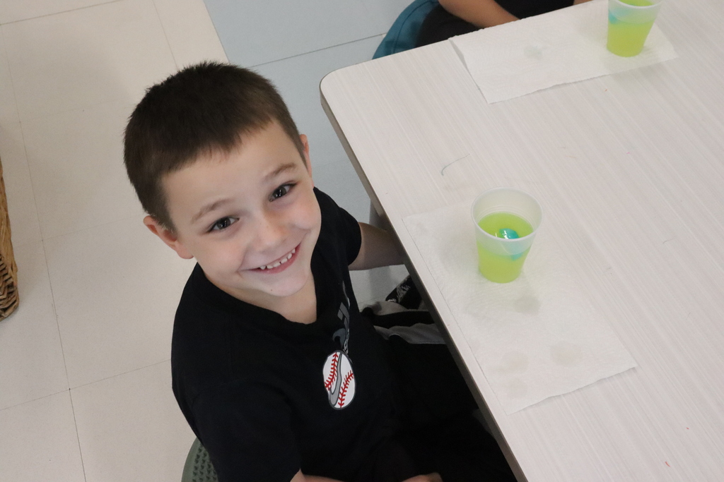 A child smiles at the camera, sitting at a table with a green drink in a cup