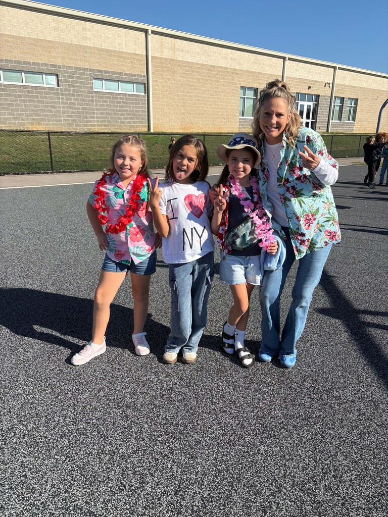 Four people stand on a playground with a school in the background. Three young girls wear leis and smile. An adult woman stands next to them wearing a tropical shirt with pink flowers.
