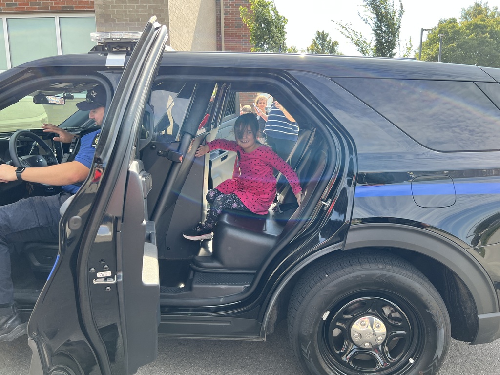 A police car with its door open. A child in a pink outfit sits inside. A police officer sits in the driver's seat.