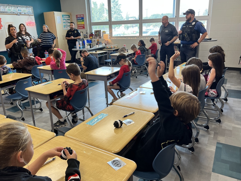 Children seated in chairs inside a classroom raising their hands, four police officers, a teacher and another adult are in the room. In the background are windows and a whiteboard.