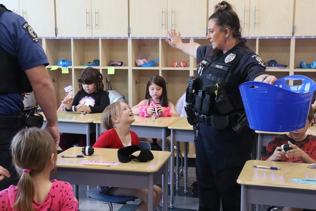 Police officer in uniform holding blue bucket, speaking to children seated at desks in classroom.