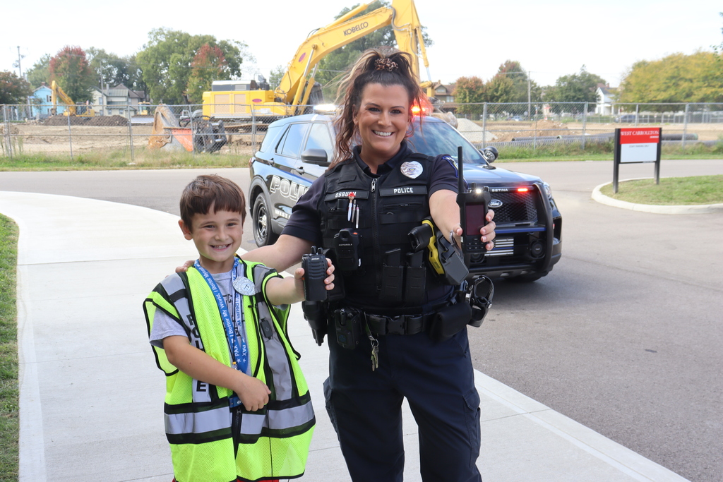 A police officer and a child in a neon green vest stand on a sidewalk, with a police car and a construction vehicle in the background.