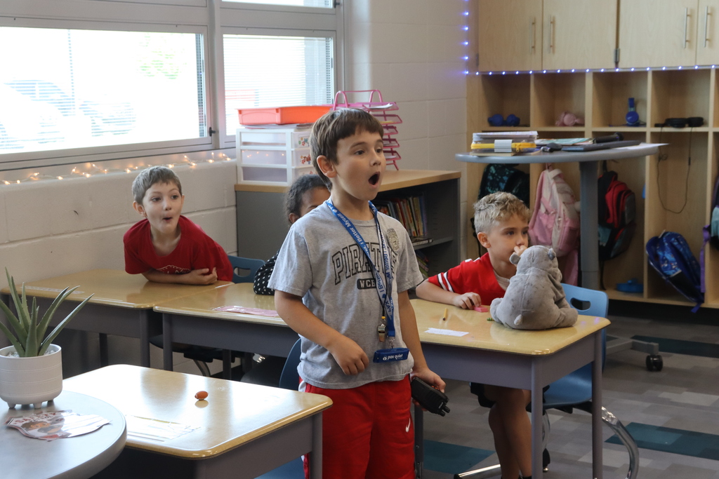 A classroom with children seated at desks; a boy stands at the front, mouth open, while others watch.